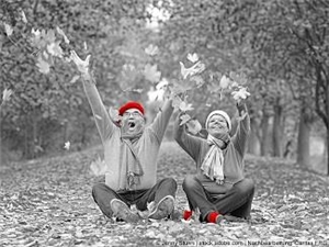 Ausgelassenes Paar sitzt im Herbstwald am Boden und wirft mit Laub. Graustufen mit roten Akzenten (Mütze, Schuhe). (c) Jenny Sturm | stock.adobe.com | Nachbearbeitung Caritas FRG.