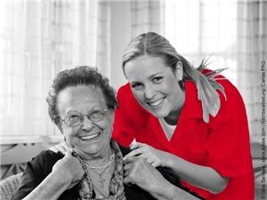 Junge Pflegerin strahlt mit einer Seniorin (sitzend) in die Kamera. In Graustufen. Shirt der Pflegekraft rot. (c) Gina Sanders|stock.adobe.com|Nachbearb. Caritas FRG.