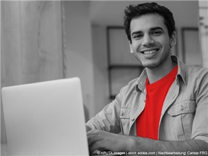 Junger Mann, mit rotem T-Shirt, lächelt hinter Laptop sitzend in die Kamera. In Graustufen. (c) ARUTA Images|stock.adobe.com|Nachbearbeitung: Caritas FRG.