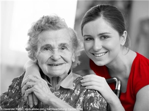 Sitzende Seniorin wird von hinten von einer jungen Frau im roten Shirt umarmt. Ansonsten in Graustufen. (c) Alexander Raths | stock.adobe.com | Nachbearbeitung Caritas FRG.