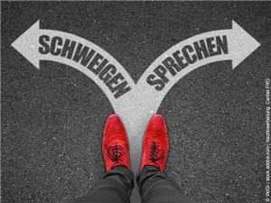 sw kolloriertes Foto. 2 Herrenfüsse in Draufsicht in roten Schnürschuhen, illustrierte Pfeile (links: "SCHWEIGEN" und lrechts: "SPRECHEN". (c) WbGi|stock.adobe.com| Nachbearbeitung: Caritas FRG.