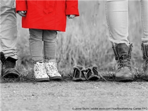 Totale auf Beine: 2 Erwachsene, in der Mitte ein Kind und ein Paar leere Kinderschuhe. In Graustufen. Kindermantel in Rot. © Jenny Sturm|stock.adobe.com|bearb.Caritas FRG