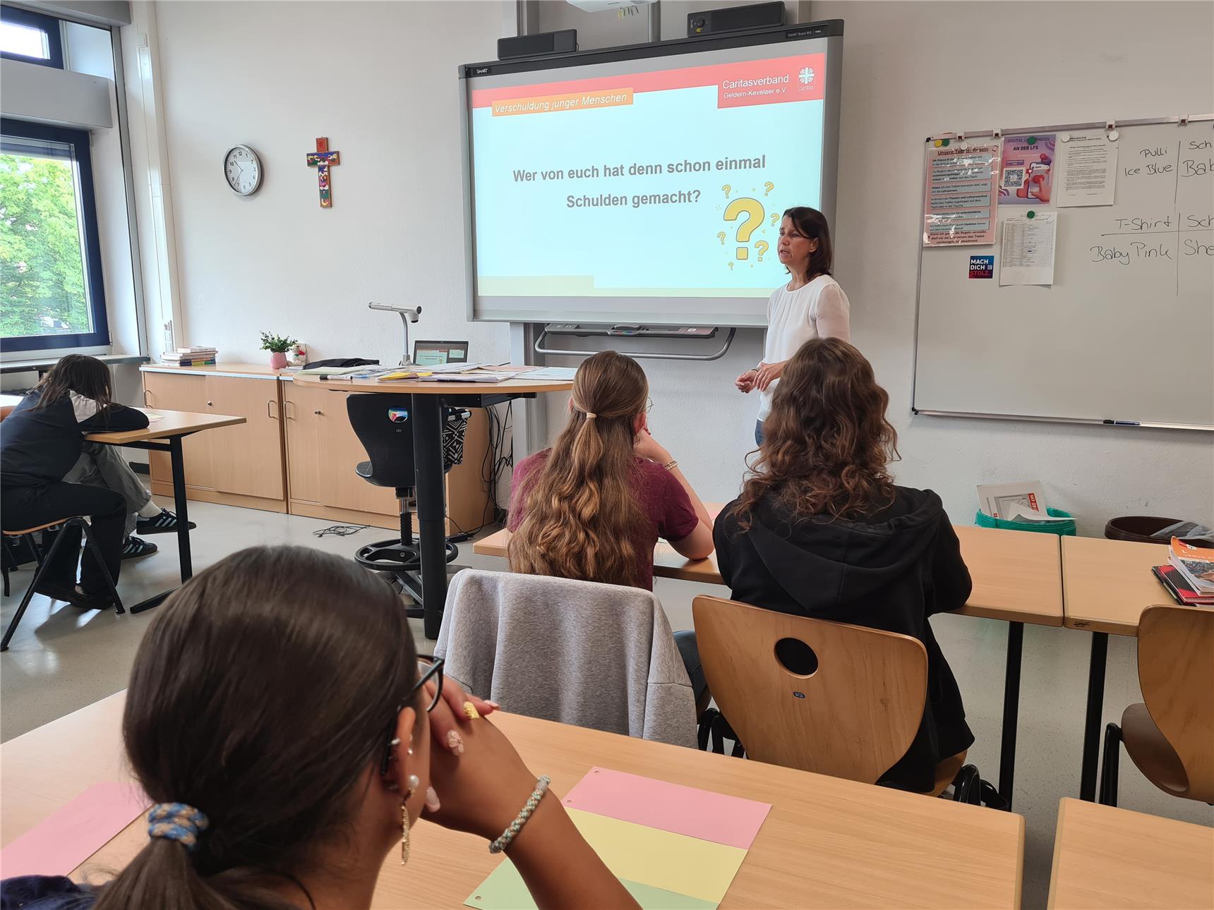Eine Mitarbeiterin der Caritas steht in einem Klassenraum einer Schule. Von hinten sind Schüler:innen an ihren Tischen sitzend erkennbar. Auf der digitalen Tafel ist der Satz 