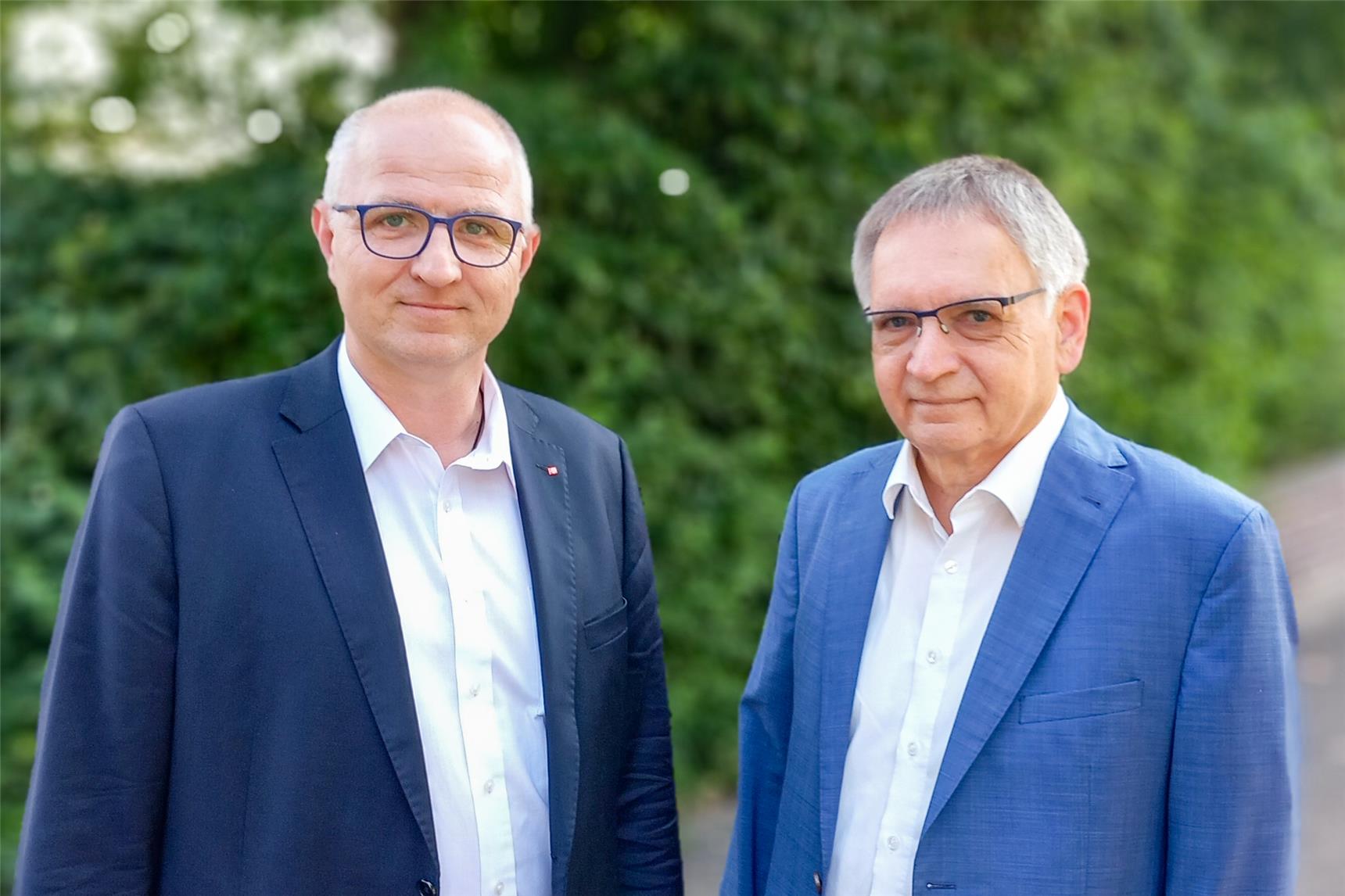 Zwei Männer in Hemd und Sakko stehen nebeneinander und lächeln in die Kamera. Im Hintergrund sind Bäume oder Sträucher zu sehen.