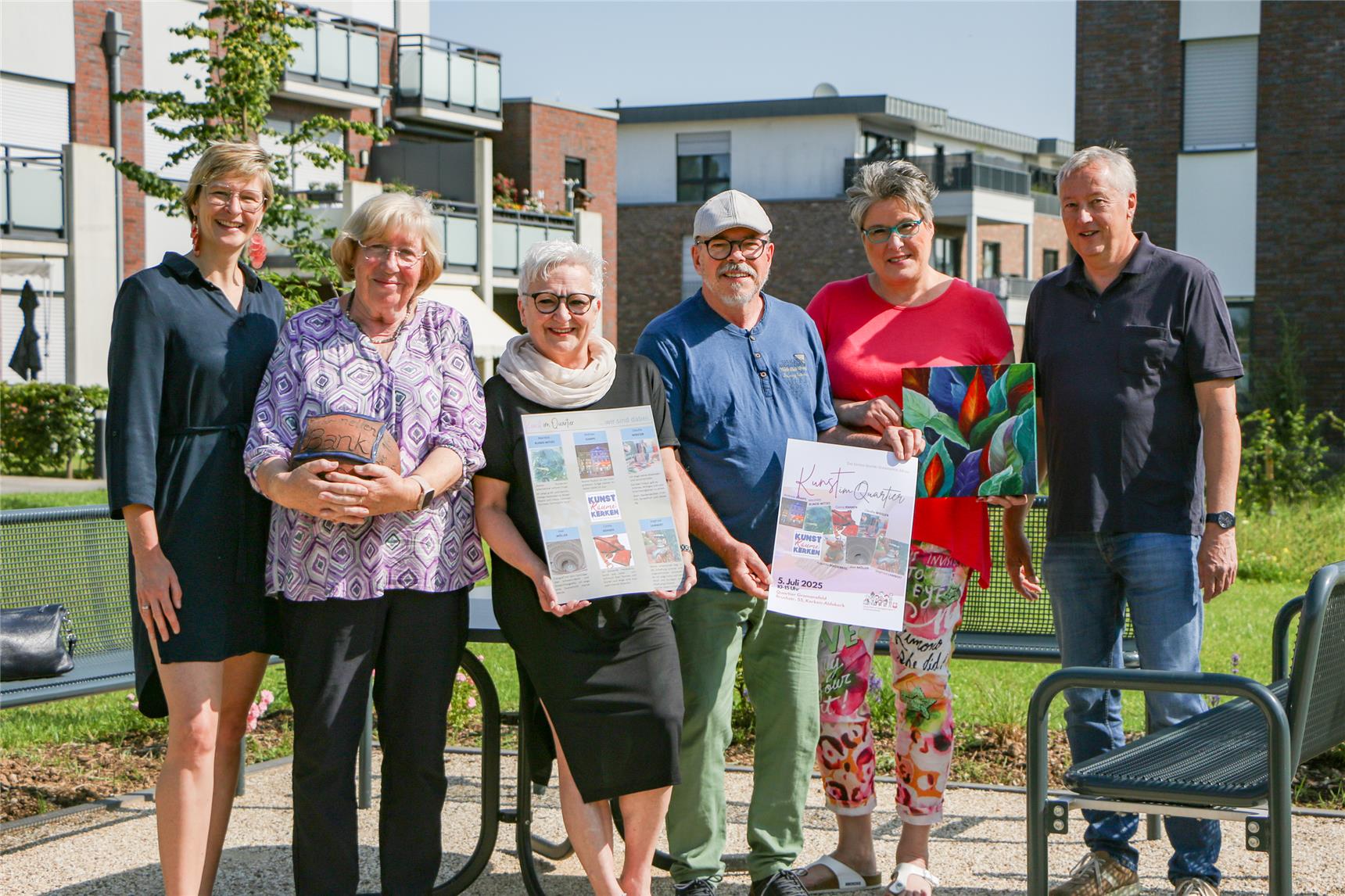 Vier weibliche und zwei männliche Personen stehen nebeneinander im Freien an einer Sitzgruppe in einer Gartenanlage eines Neubaugebietes. Einige von ihnen halten das Plakat zur Veranstaltung oder ein Kunstobjekt in den Händen.