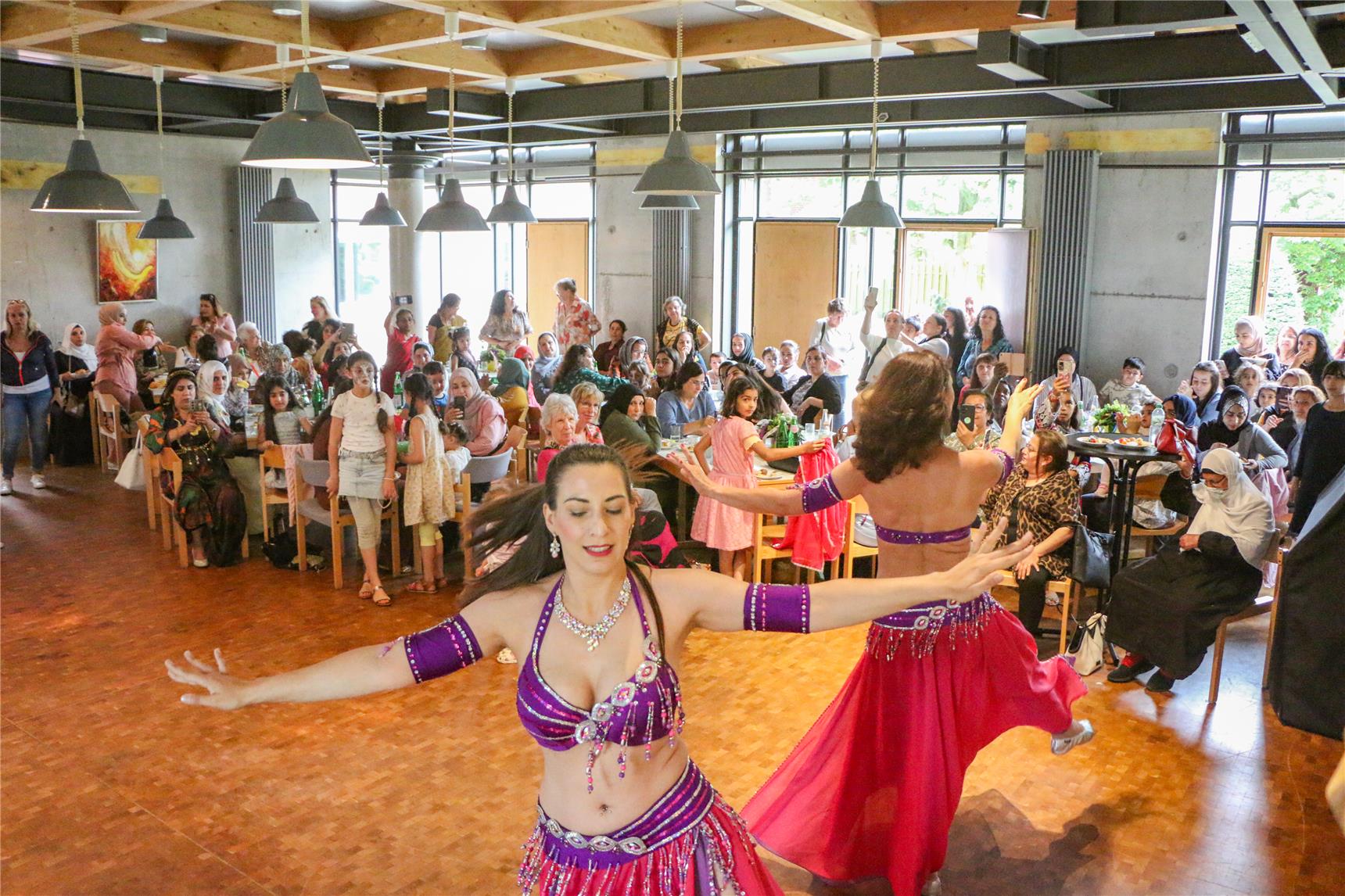 Zwei Frauen in orientalischer Tracht machen Bauchtanz in einem Veranstaltungssaal, der sehr gut gefüllt ist. Im Publikum sitzen an vielen Tischen ausschließlich Frauen und einige Kinder. Viele der Frauen tragen ein Kopftuch oder Kleidung, die auf eine nicht-europäische Herkunft schließen lassen.