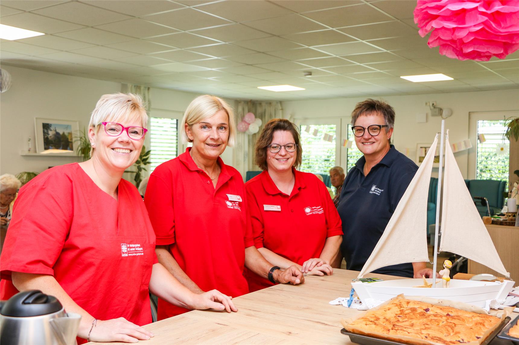 Vier Frauen stehen nebeneinander an der Anrichte einer Küche und lächeln in die Kamera. An ihrer einheitlichen Kleidung lässt sich erkennen, dass sie Mitarbeitende der Caritas sind. Im Hintergrund sind einige Senioren erkennbar.