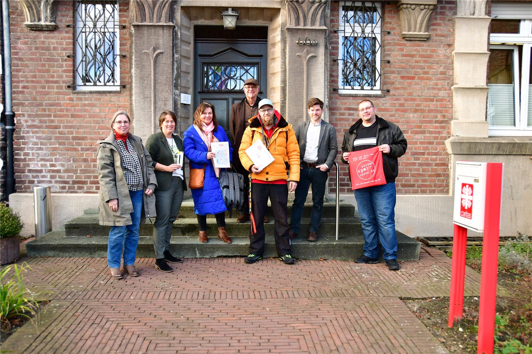 Bianka Bruckmann, Rita Fergen, Carmen Pelmter, Udo Warstat, Markus Jakobs, Gerrit Hermans und Tim Hofmeyer stehen nebeneinander vor einem Gebäudeeingang der Caritas