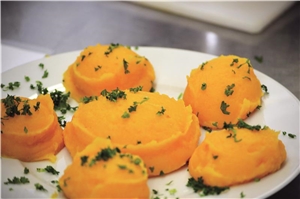 Das Bild zeigt einen Teller mit einem orange-farbenen Gemüsepüree, welches sorgfältig auf dem Teller angerichtet ist und mit Kräutern garniert wurde.