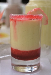 Das Bild ist eine Nahaufnahme eines Glases, welches im unteren Teil mit einem roten Mouse befüllt wurde, worauf eine helle Creme geschichtet wurde. Das Glas ist mit einem Zuckerrand dekoriert.