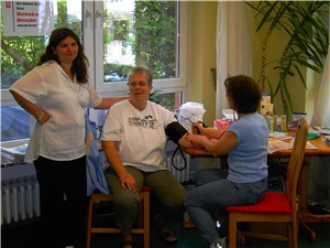 In der Mitte sitzt eine Frau auf einem Stuhl, die sich von der sitzenden Dame zu ihrer rechten den Blutdruck messen lässt. Zu ihrer linken steht eine Frau, die ihren Arm auf ihre Schultern legt.
