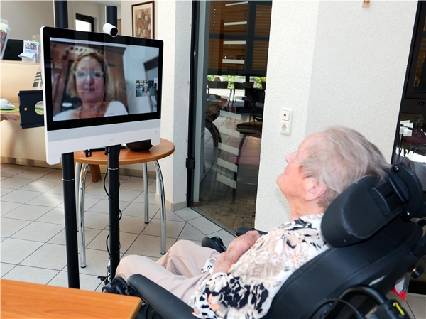 Eine alte Frau sitzt im Rollstuhl vor einem Bildschirm, auf dem eine Frau zu sehen ist.