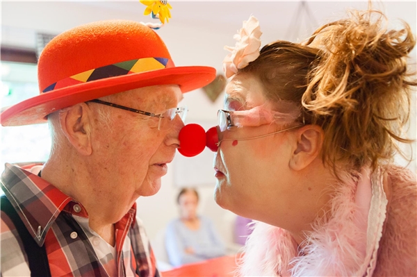 Eine Clownin berühr mit der roten Clownnase einen alten Mann, der ebenfalls eine rote Nase trägt.