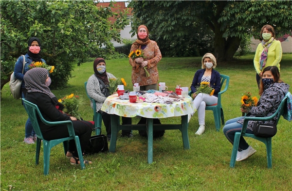 Frauen mit Masken und Blumen um einen Tisch, auf dem weitere Masken liegen.