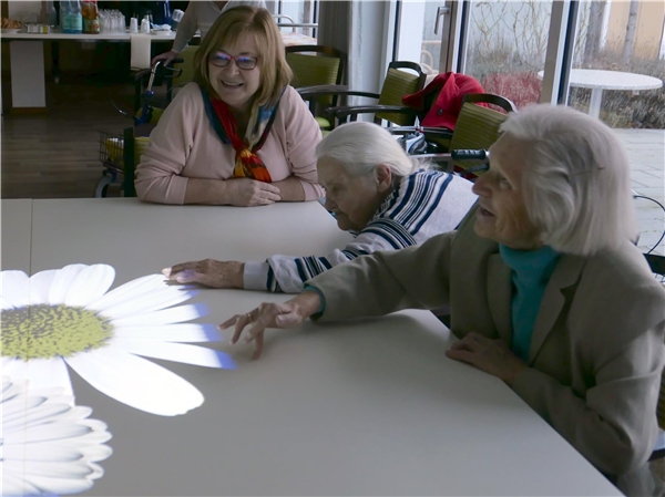 Drei Frauen berühren eine Lichtprojektion in Form einer Blume auf einem Tisch.