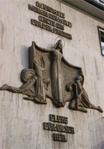 Maria Frieden Figuren einer Frau und zweier Männer an einer Hauswand. Die Frau hat einen Heiligenschein und breitet die Arme über den knienden Männern aus.