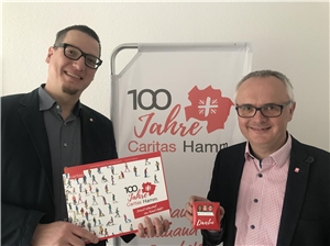 Zwei Männer halten zeigen Broschüre 100 Jahre Caritas Hamm und eine Pralinenpackung mit "Danke" 
