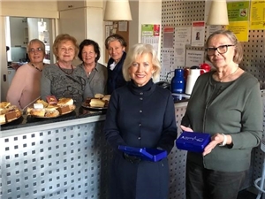 Zwei Frauen stehen vor der Kuchentheke mit blauen Brotdosen in der Hand, vier Frauen stehen hinter der Theke