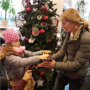 Vor einem geschmückten Weihnachtsbaum übergibt ein kleines Mädchen eine Spendendose an eine Frau