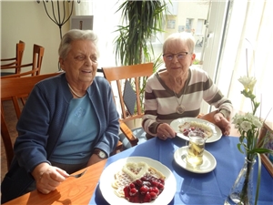 Zwei Seniorinnen sitzen am Tisch vor je einem Teller mit einer Herzchenwaffel mit Kirschen und Sahne