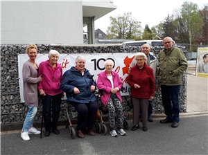 Sechs Senioren stehen oder sitzen auf ihren Rollatoren vor einem Plakat "100 Jahre Caritas Hamm"