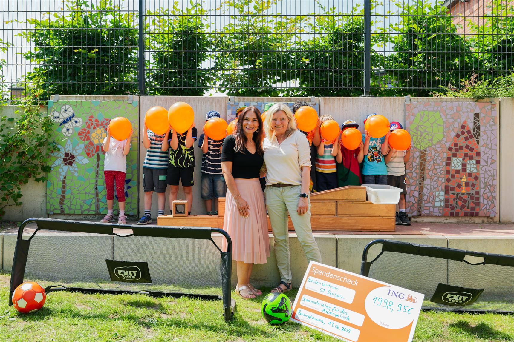 Martha Suszyk und Angela Boyksen stehen neben dem Spendencheck und den Fußballtoren. Im Hintergrund stehen einige Kinder der Einrichtung mit Luftballons vor dem Gesicht. 