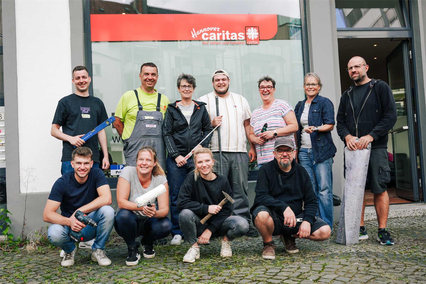 Mehrere Reemtsma-Mitarbeitende sowie Ramona Ligendza und David Ohnesorge von der Caritas stehen mit Werkzeug in der Hand vor dem Gebäude des Kontakt Cafés. Sie blicken in die Kamera und lächeln.