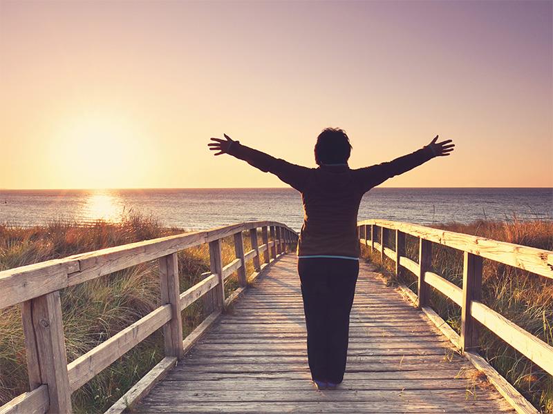 Eine Frau, die von hinten zu sehen ist, steht mit ausgebreiteten Armen auf einem Holzsteg in den Dünen, mit Blick auf den Strand, die Ostsee und den Sonnenaufgang.