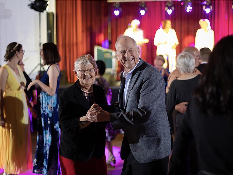 Ein Seniorenpaar tanzt Tango und lacht dabei in die Kamera. Im Hintergrund tanzen und unterhalten sich weitere Menschen in Abendgarderobe.