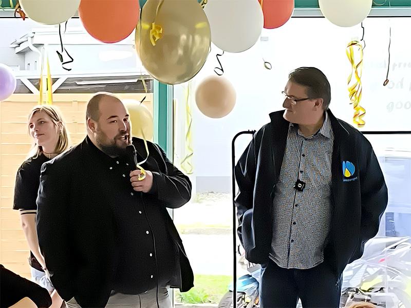Zwei Männer stehen nebeneinander in einem Raum, der linke hält ein Mikrofon, über ihnen hängen bunte Luftballons