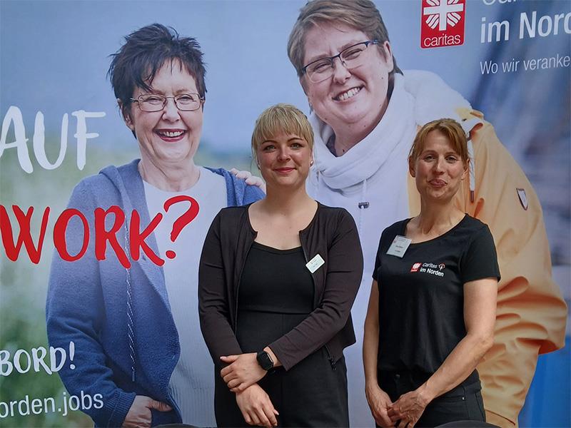 Zwei Frauen stehen vor einer Plakatwand mit einem Plakat der Caritas, auf dem zwei andere Frauen abgebildet sind.