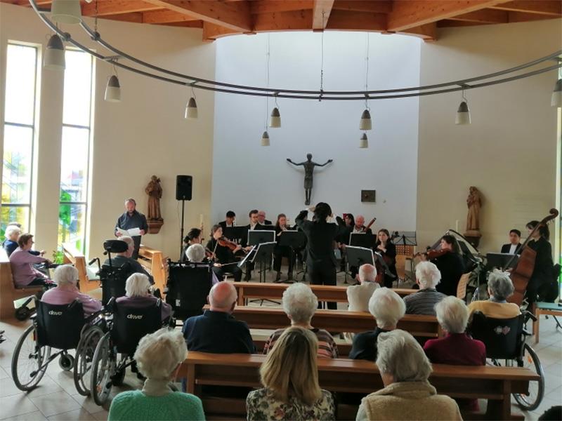 Seniorinnen und Senioren sitzen in einer Kapelle und hören einem Orcchester zu; 