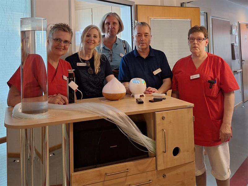 Vier Frauen, davon zwei in roter Dienstkleidung, und ein Mann stehen hinter einem Wagen aus Holz miz Schubladen, einer wassergefüllten Glassäule, Lichterkabeln und anderen Hilfsmitteln. 