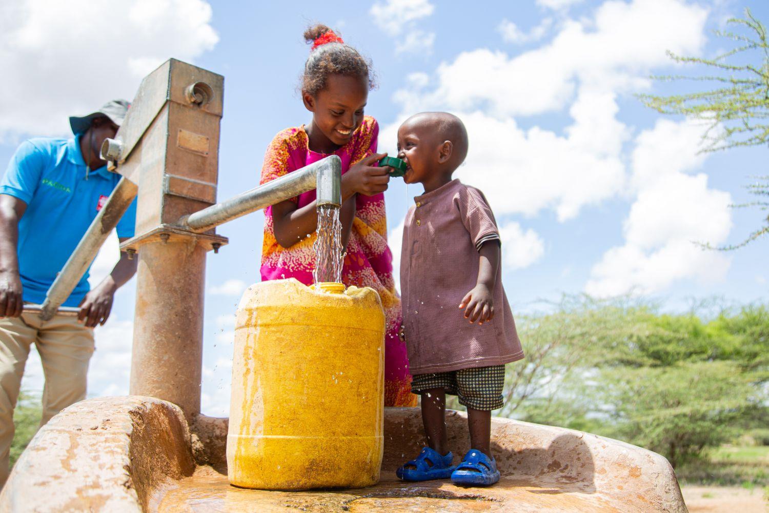 Adho hilft ihrer kleinen Schwester beim Trinken an einem Brunnen in Kenia. PACIDA hilft, den Wassernotstand in Afrika zu überwinden.