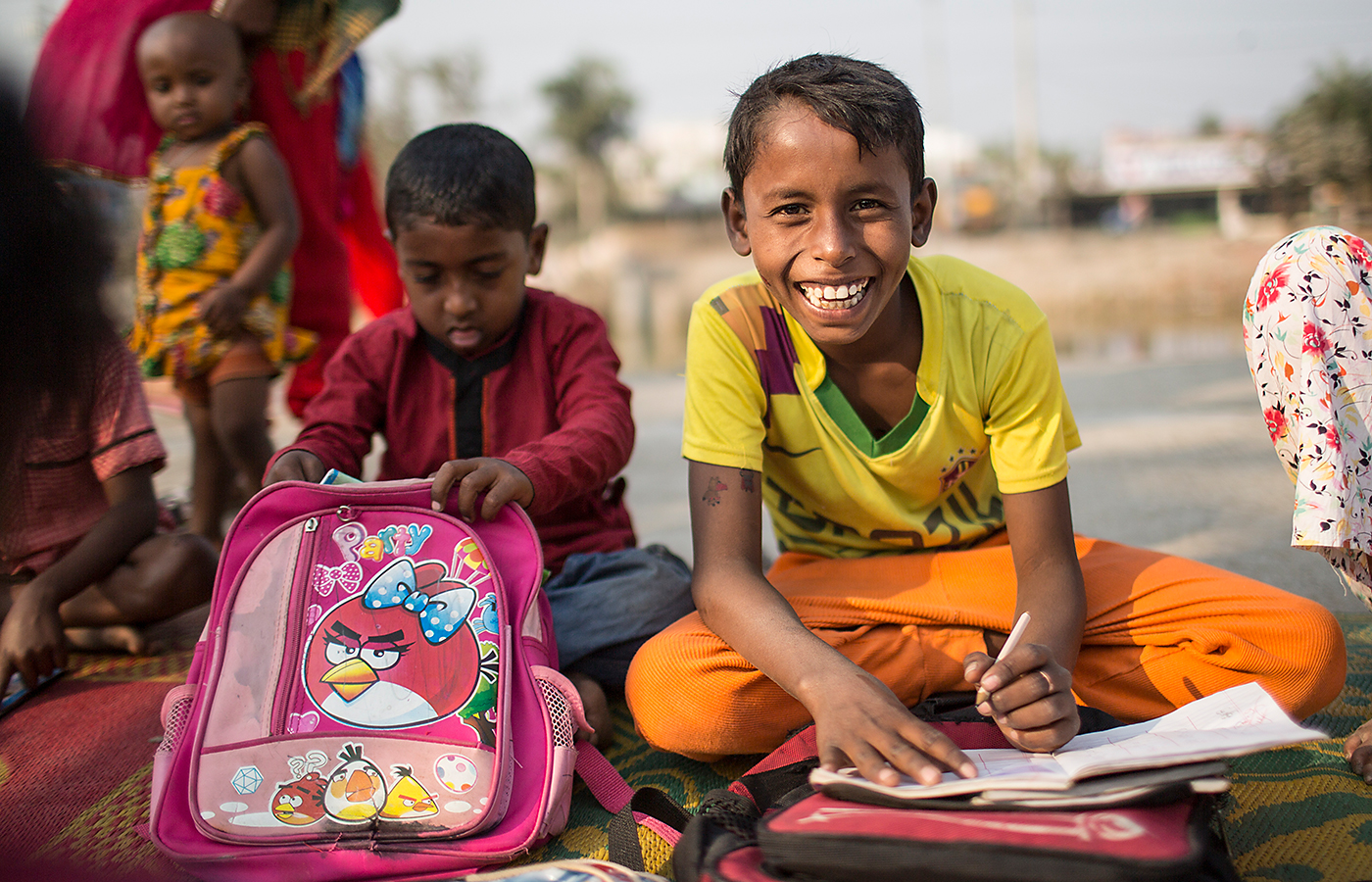Lachender Junge schreibt in ein Schulheft – Open-Air-Schule in Rajshahi / Bangladesch