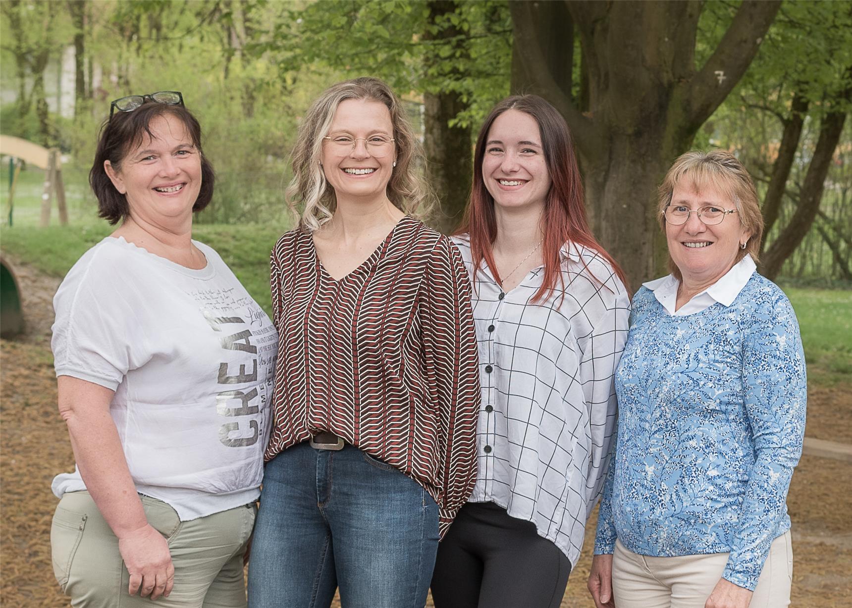 Vier Frauen stehen gemeinsam vor Bäumen und lächeln, umgeben von einer natürlichen Umgebung.