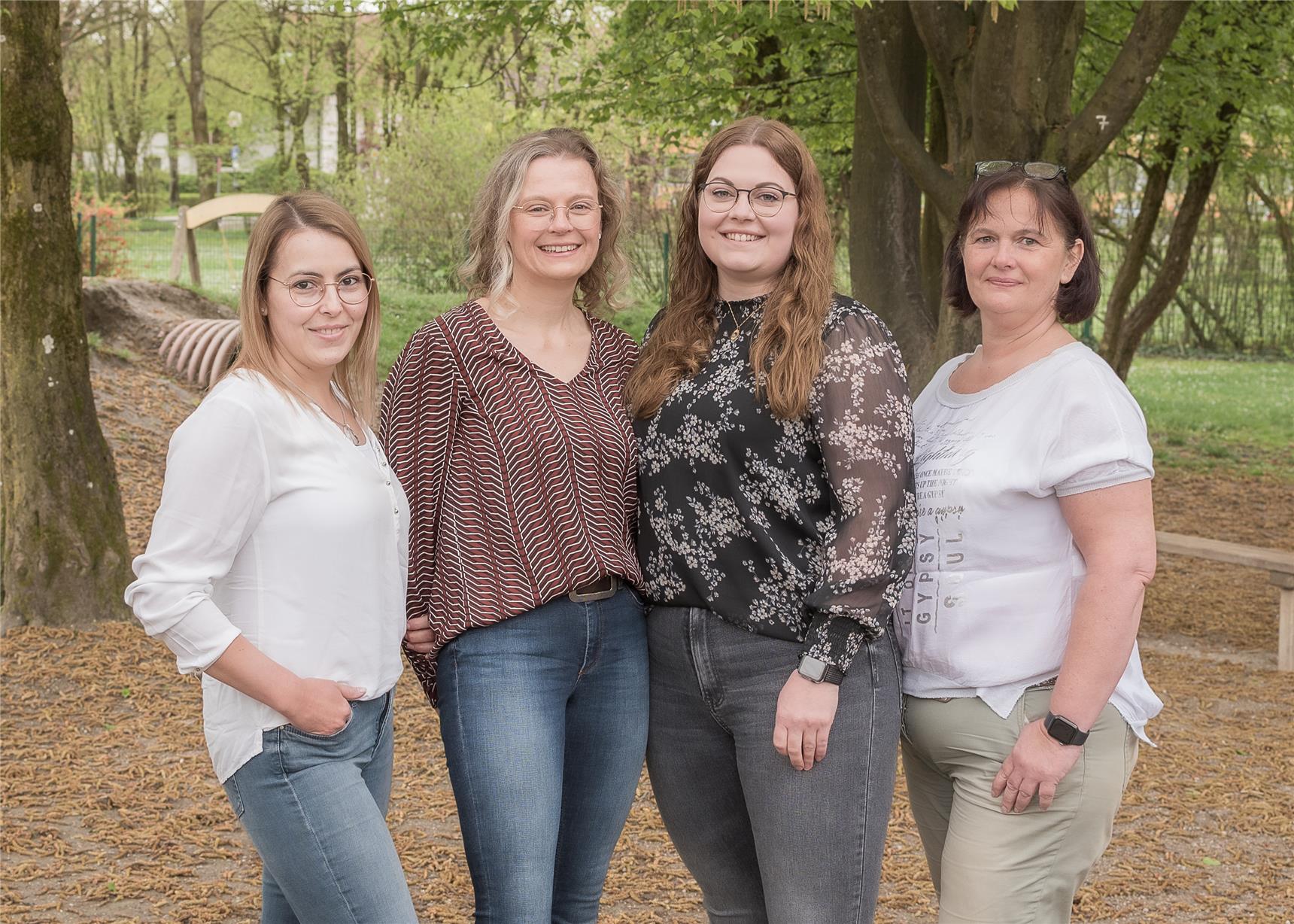 Vier Frauen posieren lächelnd vor einer Baumgruppe in einer grünen Landschaft.