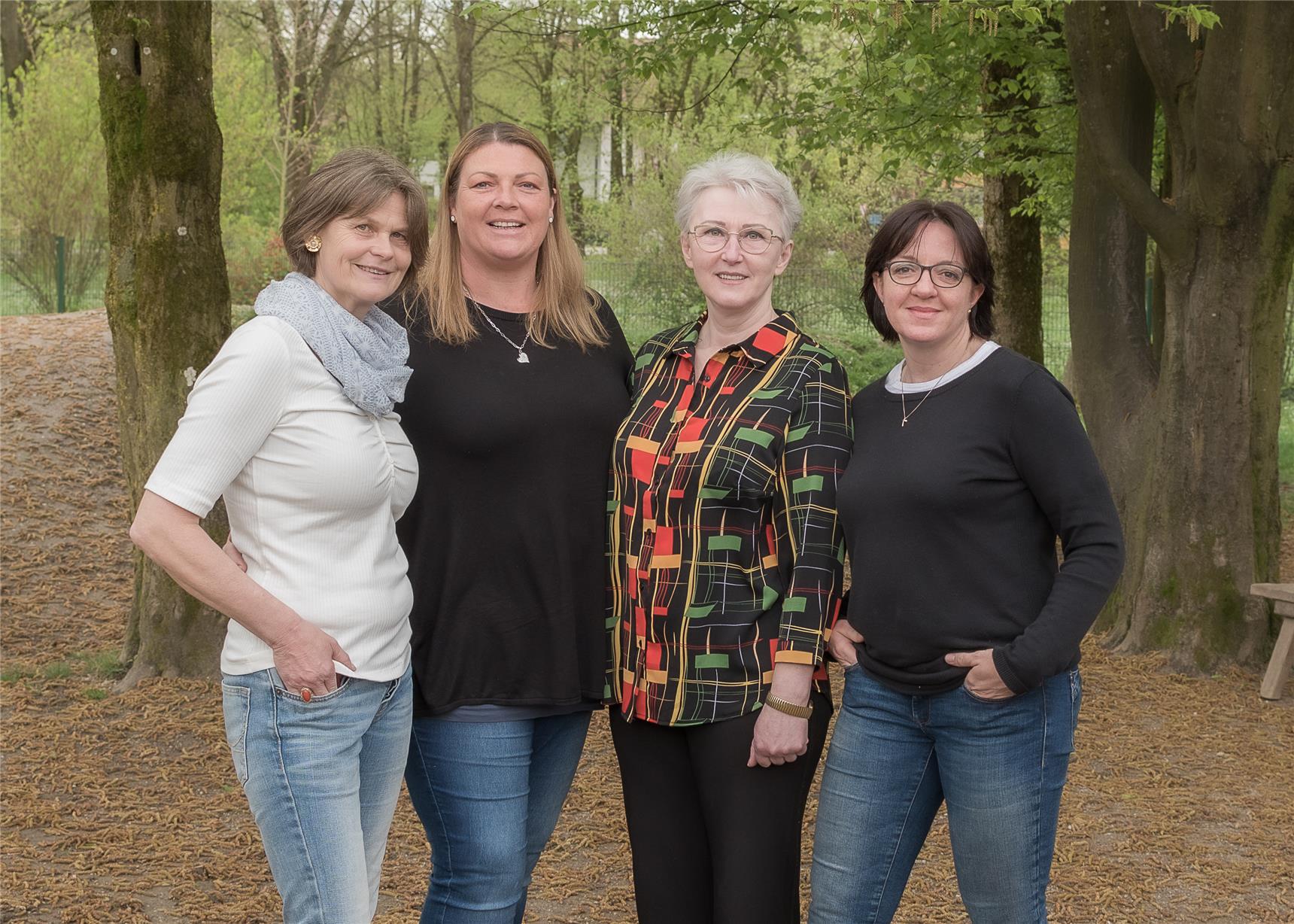 Vier Frauen stehen vor Bäumen und lächeln, umgeben von einer natürlichen Umgebung.