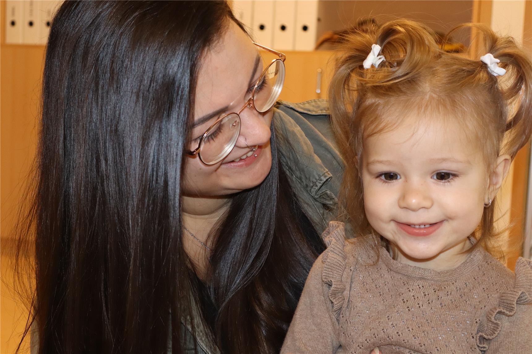 Eine junge Frau mit Brille und langen dunklen Haaren lächelt das fröhliche kleine Mädchen neben ihr an. Das Kind trägt einen braunen Pullover und hat zwei verspielte Zöpfe mit weißen Schleifen.
