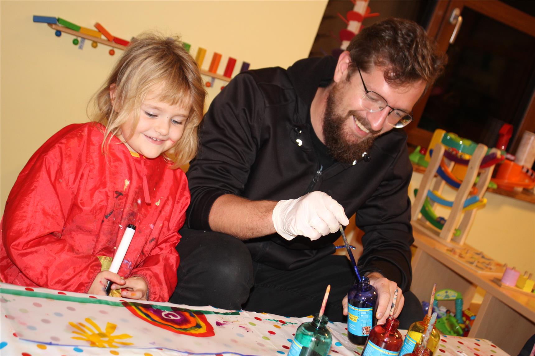 Ein Mann mit Brille und Bart malt lachend mit einem Kind, das eine rote Malschürze trägt. Vor ihnen liegen bunte Farben, Pinsel und ein fröhlich gestaltetes Bild.