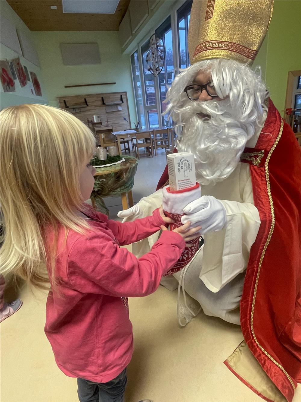 in als Nikolaus verkleideter Mann mit rotem Umhang und goldener Mitra überreicht einem kleinen Mädchen in pinkem Pullover ein Geschenk. Die Szene spielt sich in einem gemütlich dekorierten Raum ab.