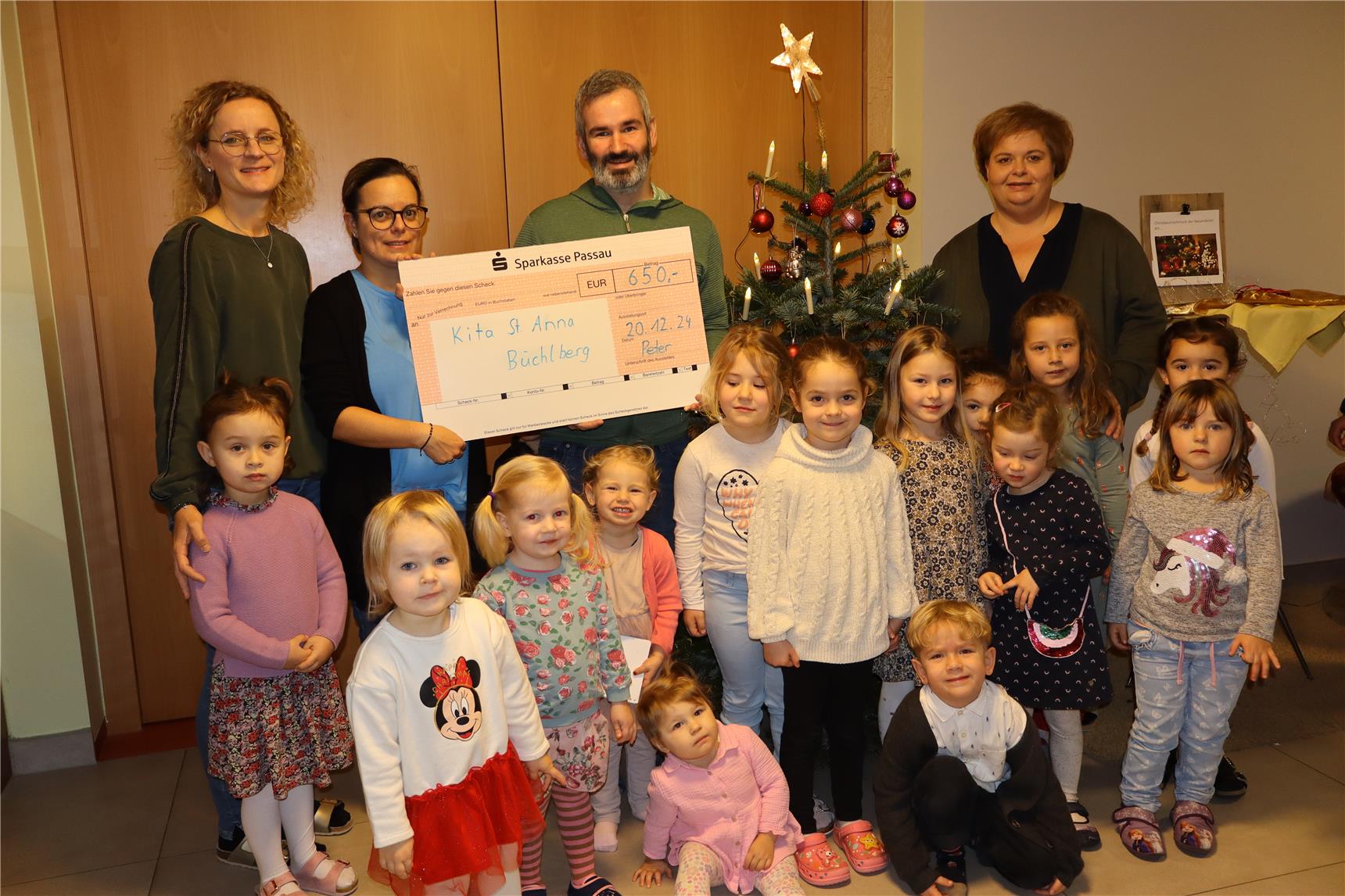 Eine Gruppe von Kindern und Erwachsenen präsentiert eine Spende für die Kita St. Anna Büchlberg. Sie stehen vor einem geschmückten Weihnachtsbaum.