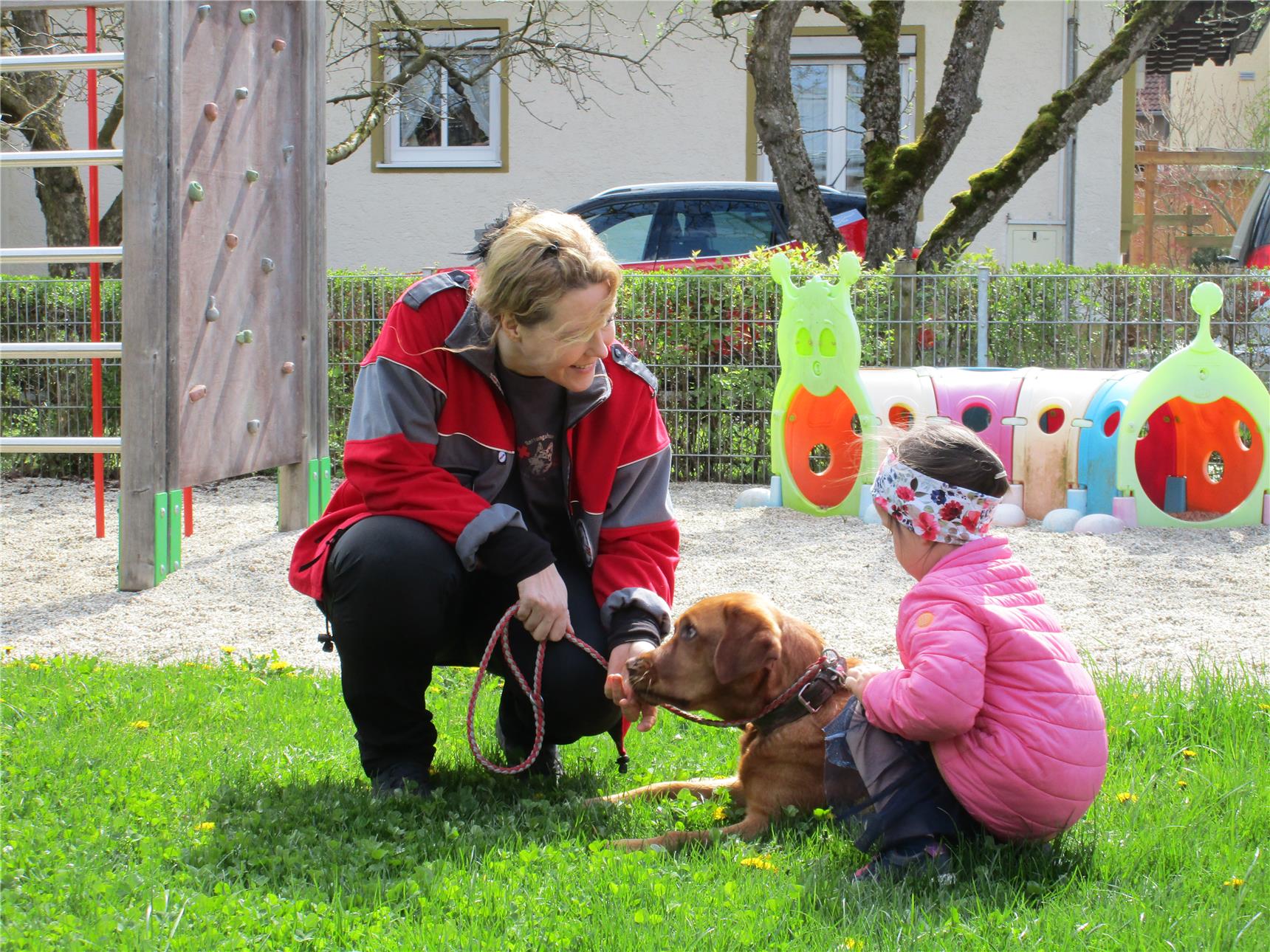 Zwei Personen, eine erwachsene Frau in roter Jacke und ein kleines Kind in rosa Jacke und Stirnband, sitzen auf einer Wiese neben einem braunen Hund. Die Frau hält die Leine, während das Kind den Hund sanft streichelt; im Hintergrund ist ein Spielplatz mit buntem Klettertunnel und Kletterwand zu sehen.