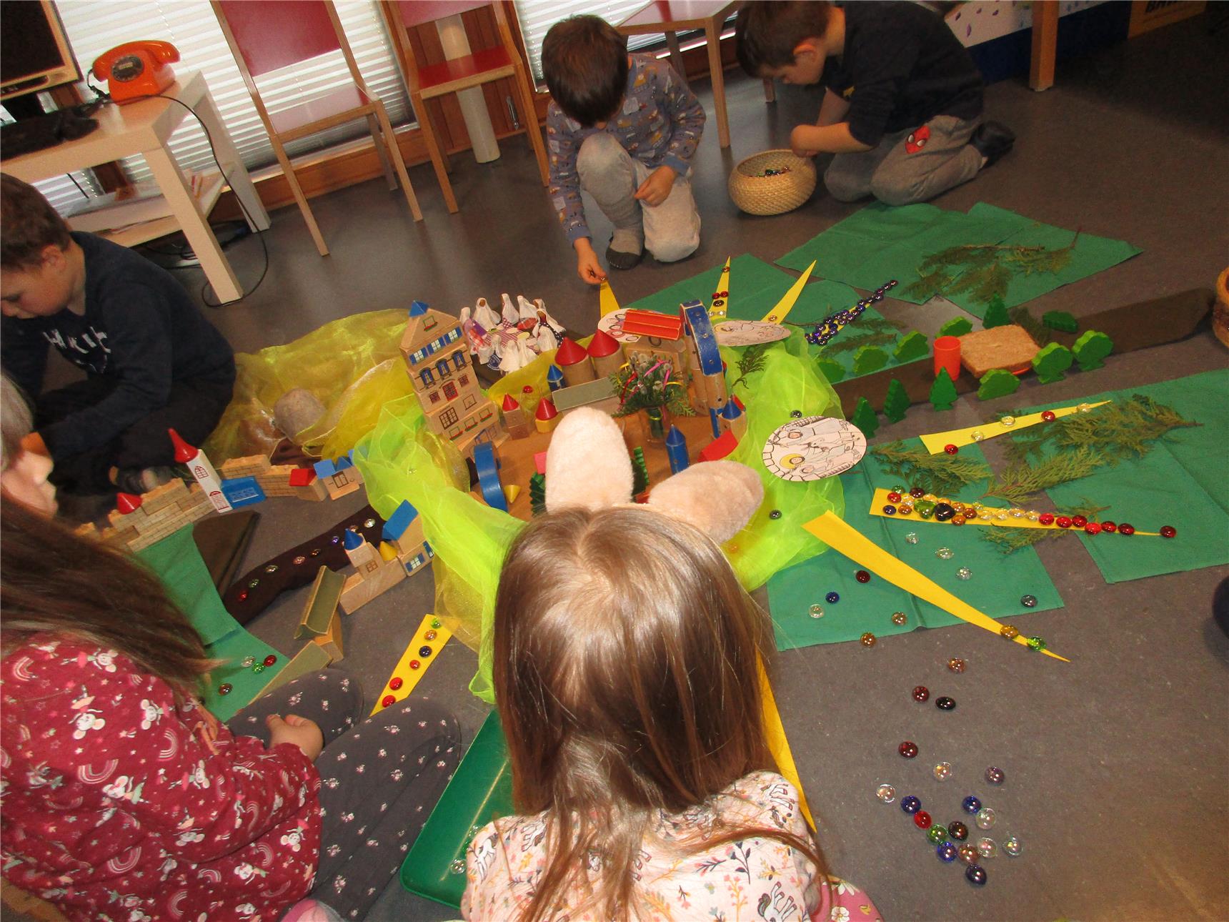 Mehrere Kinder sitzen auf dem Boden in einem Stuhlkreis und gestalten gemeinsam eine bunte Osterlandschaft mit Bausteinen, Figuren, Naturmaterialien, Glassteinen und bemalten Bildern. In der Mitte steht eine symbolische Stadt aus Holzklötzen, gelben Strahlen und grünem Tuch – vermutlich Teil einer religiösen Osterfeier oder eines Projekttags im Kindergarten.