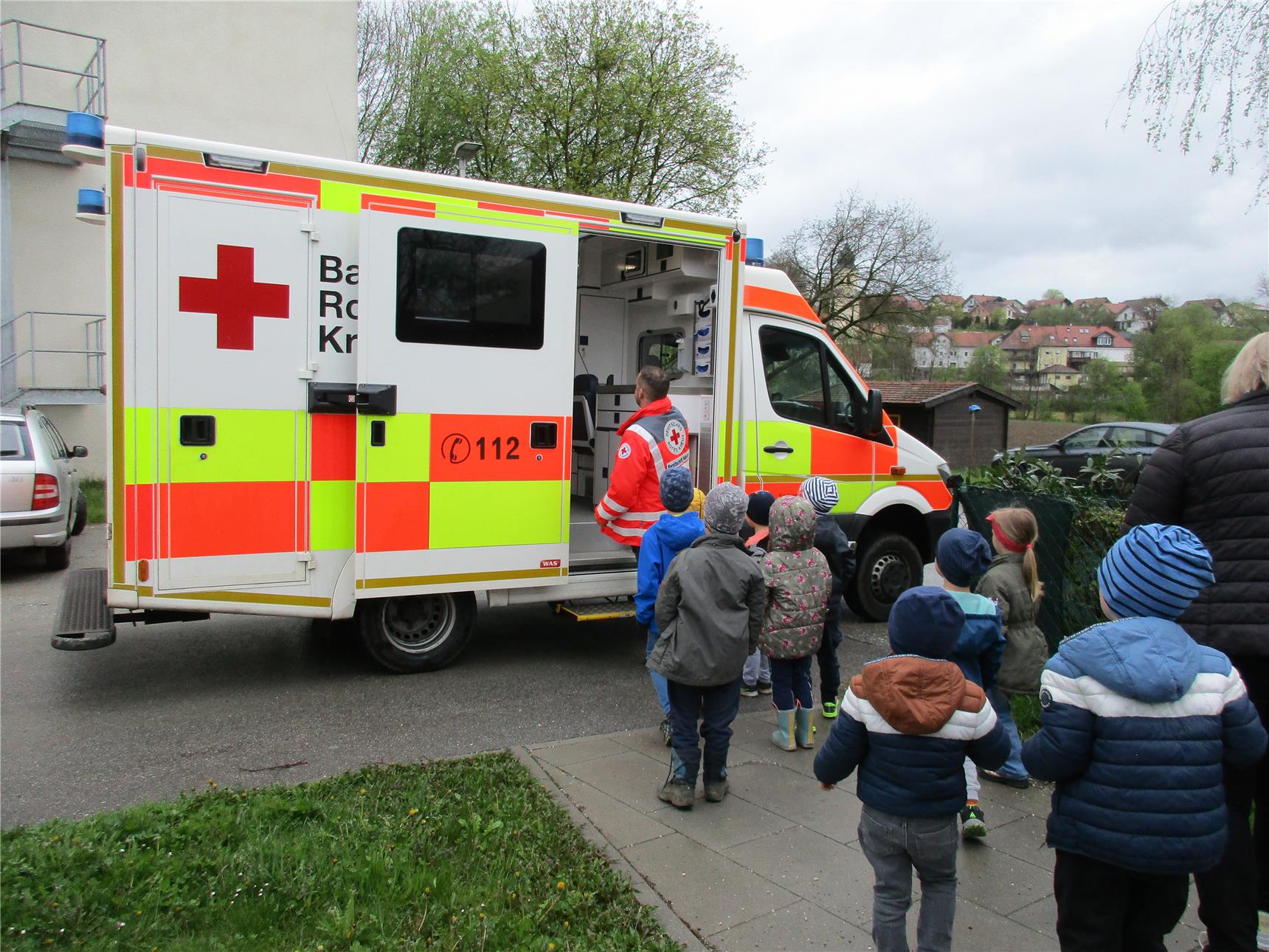 Eine Gruppe Kindergartenkinder schaut sich gemeinsam mit einer Erzieherin einen Rettungswagen des Bayerischen Roten Kreuzes an, dessen Seitentür geöffnet ist. Ein Sanitäter in roter Einsatzkleidung erklärt den Kindern das Fahrzeug, das am Straßenrand vor einem Gebäude steht.