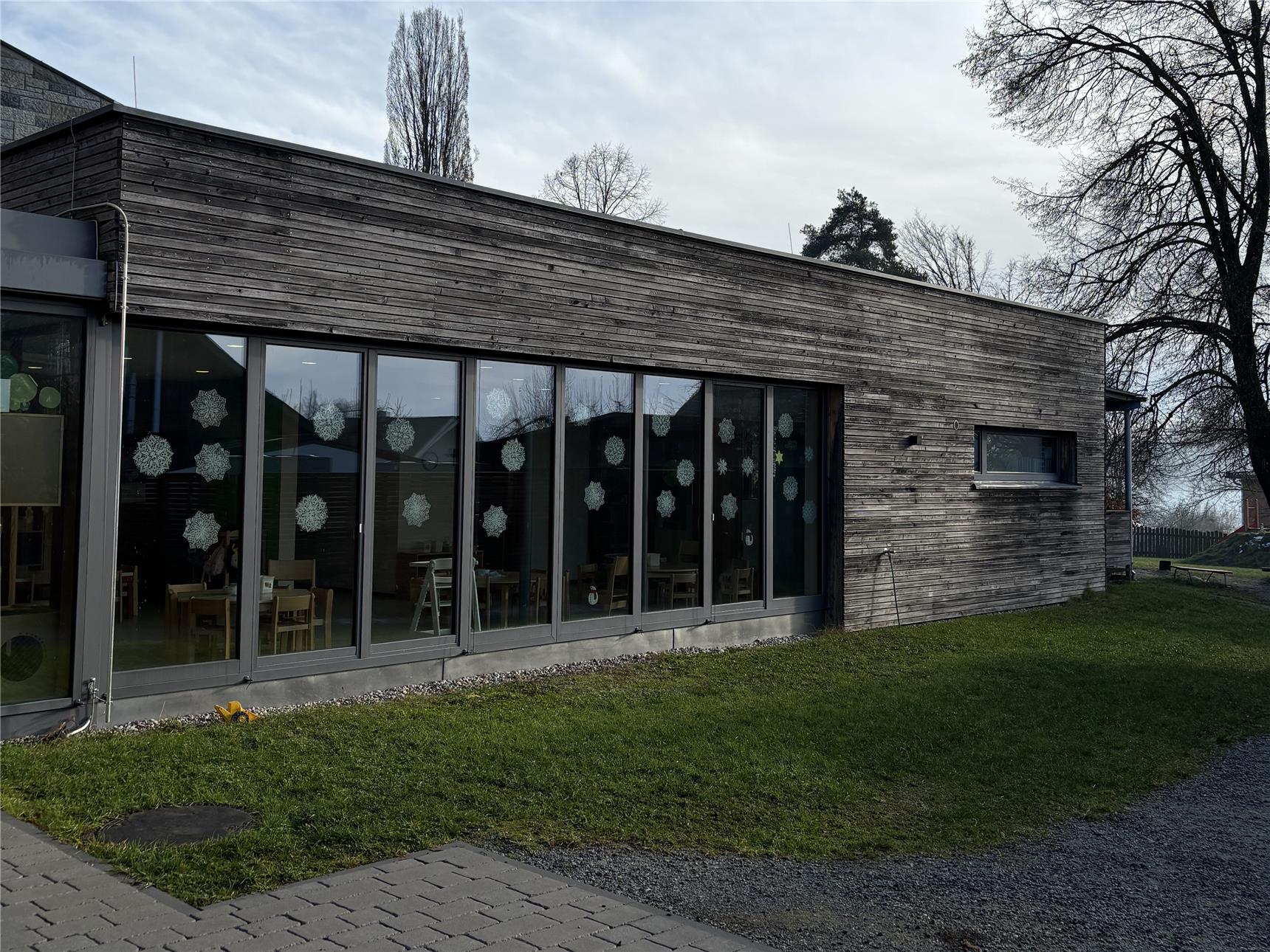 Ein eingeschossiges Gebäude mit Holzverkleidung und großen Fenstern, durch die ein Innenraum mit Tischen und Stühlen sichtbar ist. Vor dem Gebäude erstreckt sich eine Grünfläche.