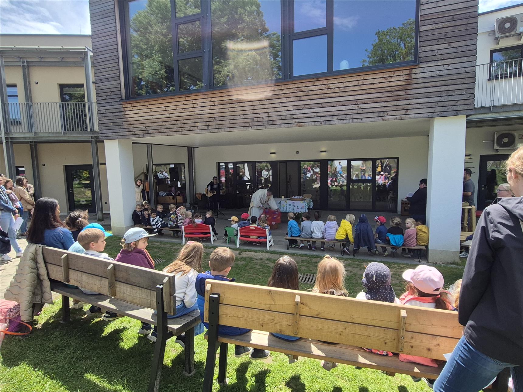 Eine Gruppe von Kindern sitzt draußen auf Bänken und kleinen Stühlen vor einem modernen Schul- oder Kindergarten-Gebäude mit Holzverkleidung und großen Fenstern. Im Hintergrund findet offenbar eine Aufführung oder Veranstaltung statt – eine Frau steht an einem Tisch mit einem Tischtuch, während Erwachsene im Hintergrund musizieren und andere zuschauen. Es scheint ein sonniger Tag mit fröhlicher Stimmung zu sein