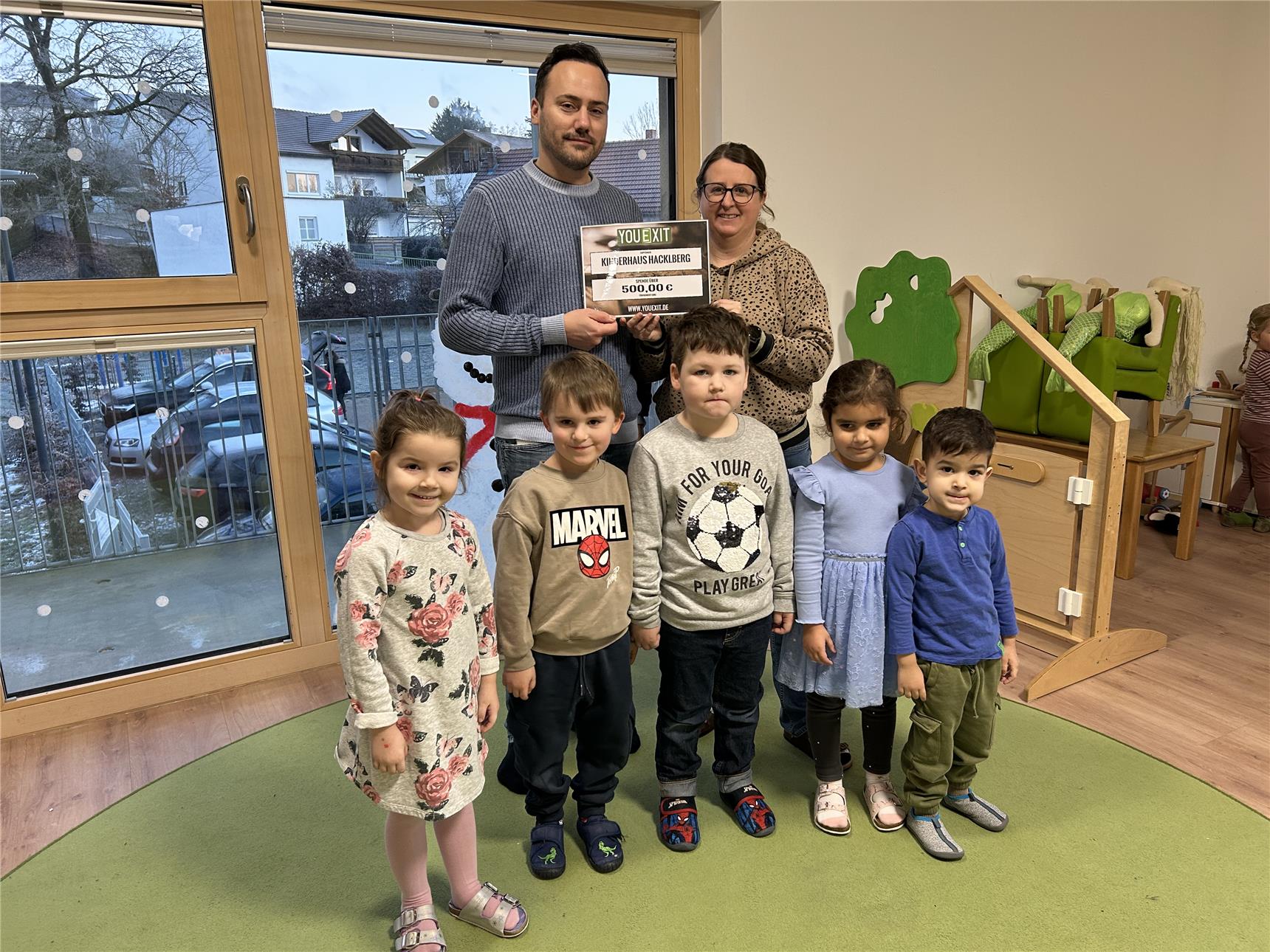 Ein Mann und eine Frau halten eine Spendentafel mit der Aufschrift „YOU EXIT“ und „500,00 €“ für die Kita Haus Hacklberg, umgeben von sechs fröhlichen Kindern.