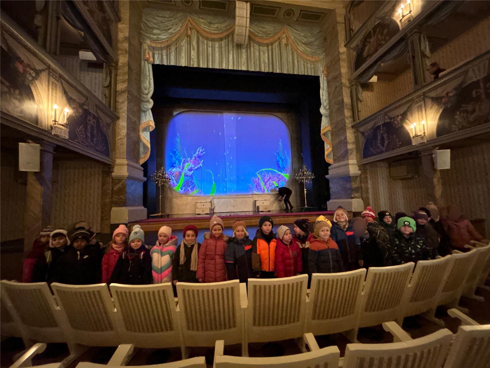 Eine Gruppe von Kindern in bunter Winterkleidung steht in einem prunkvollen Theater und blickt zur Kamera. Die Bühne im Hintergrund zeigt ein blau beleuchtetes Unterwasser-Motiv.