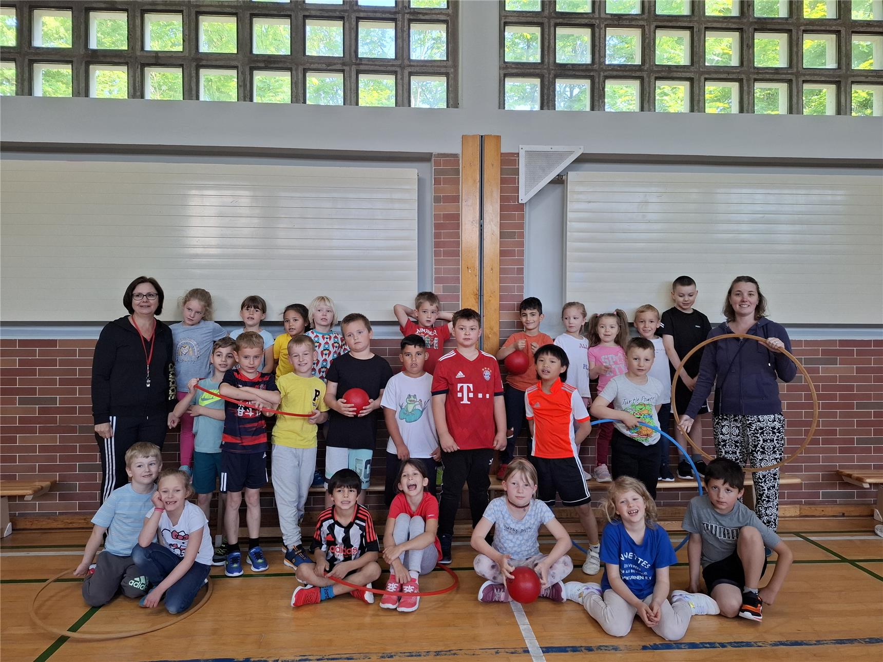 Gruppenfoto von Kindern und zwei Lehrerinnen in einer Turnhalle, umgeben von Sportgeräten wie Bällen und Reifen. Die Kinder lachen und posieren in Sportkleidung nach einer Turnstunde.